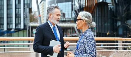 Two people stand facing each other in conversation on an outdoor terrace with a modern city building behind them. One wears a dark suit and tie and holds a tablet; the other wears a patterned blue top. Both appear engaged in discussion.