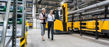 Two people walk side by side through a large industrial facility, one holding a tablet and the other gesturing while speaking. Yellow heavy machinery and industrial shelving are visible in the background.