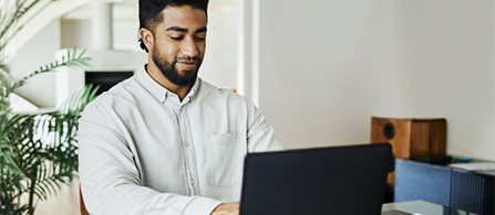 A person with a beard wearing a light gray shirt sits at a desk in a bright, airy home or modern office, focused on typing on a laptop. A plant and speaker are visible in the background.