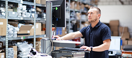A person wearing a dark navy polo shirt stands at a mobile workstation with a mounted monitor in an industrial warehouse or parts storage facility, focused on the screen. Shelves with small components are visible in the background.