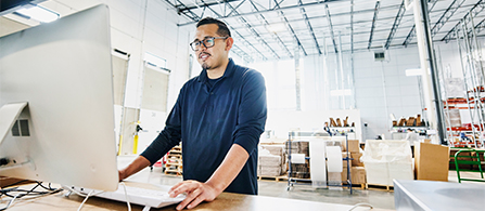 A person wearing glasses and a dark long-sleeve shirt stands at a desktop computer in a bright warehouse or logistics facility, looking at the monitor with a focused expression. Boxed inventory and shelving extend into the background.