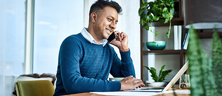 A person wearing a blue sweater sits at a desk, smiling while talking on a phone and looking at an open laptop. Greenery and natural light are visible through a window in the background.
