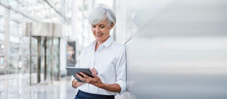A person wearing a white button-down shirt leans against a wall in a bright, modern lobby or office corridor, smiling and looking down at a tablet held in both hands. Large windows create a soft, airy atmosphere.