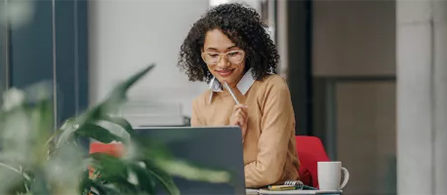 A person with curly natural hair wearing a dark teal blouse stands smiling in the foreground of a bright, modern open office, holding a tablet. Two other people are visible in conversation at tables in the background.