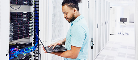 A person with dark hair and a short beard, wearing a light blue polo shirt, stands in a data center holding an open laptop on one arm and typing with the other hand while looking down at the screen. To the left is an open server rack with bundles of colorful network cables; rows of equipment racks recede into the background.
