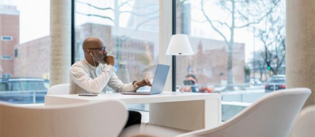 A person with a shaved head, wearing a white long-sleeved shirt, sits at a white table in a bright office with floor-to-ceiling windows. They rest their chin on one hand and look toward an open laptop, with a small lamp and a notebook on the table and empty white chairs nearby. A city view is visible through the windows.