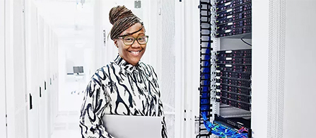 A person with their hair in a bun and wearing glasses and a black and white patterned blouse stands in a data center aisle, holding a laptop and smiling toward the camera. A server rack with small blue indicator lights and cabling is beside them, with a bright white corridor of racks behind.
