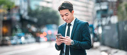 A person with short dark hair, wearing a dark blue business suit, white shirt, and tie, stands on a city street and looks down with a slight smile at a smartphone held in both hands. The street behind them is blurred, with out-of-focus traffic and buildings.
