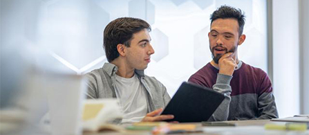 Two people sit side by side at a table in a bright office, looking together at a tablet held by one of them. One wears a gray sweater; the other wears a maroon and gray shirt and rests a hand on the chin in a thoughtful pose. Blurred paper models sit in the foreground.