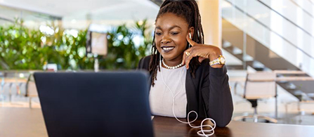 A person sits at a table in a bright, plant-filled lobby, smiling while looking at an open laptop. They wear a dark blazer over a white top and earbuds, with one hand raised near the face. Blurred greenery and lounge seating fill the background.