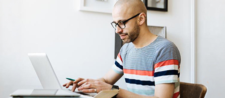 A person wearing glasses and a striped short-sleeve shirt in red, navy, and gray sits at a desk in a bright room, focused on a laptop while holding a small card in one hand. Framed pictures hang on the white wall behind them.