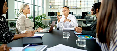 A group of people sit around a dark conference table in a modern office, engaged in discussion. One person in a white shirt gestures while speaking; the others listen. Laptops, papers, water glasses, and a tablet rest on the table.