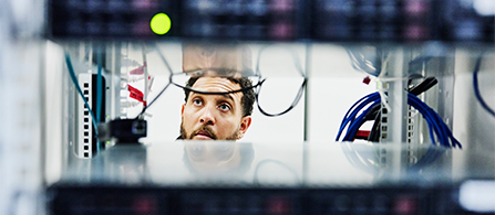 A person with a beard and glasses peers through a narrow gap between two equipment racks in a data center, surrounded by colorful network cables in red, blue, and white. A small green status light glows near the top of the frame.