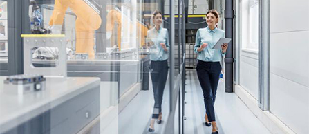 A person in a light blue blouse and dark trousers walks down a bright industrial corridor holding a tablet, passing a glass partition that reflects their image. A yellow robotic arm is visible behind the glass.