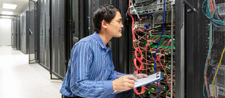 A person wearing glasses and a blue striped shirt crouches beside a server rack in a data center aisle, holding a clipboard and examining bundles of network cables in red, green, and blue. Rows of dark server cabinets recede into the background.