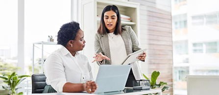 Two people work together in a bright office. One sits at a desk with a laptop, wearing a white blouse; the other stands beside them holding a document and gesturing while they talk. Large windows and greenery appear in the background.