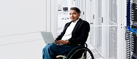 A person wearing a dark blazer over a white top is seated with a laptop in a bright white data center aisle lined with server cabinets, smiling toward the camera.