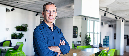 A person wearing glasses and a dark blue button-up shirt stands with arms crossed in a bright, open-plan office, looking at the camera with a slight smile. Green chairs, plants, and industrial ceiling fixtures fill the background.