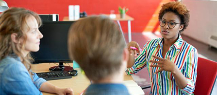 A person wearing glasses and a brightly striped shirt sits gesturing with both hands while talking to two colleagues seen from behind. A red wall and a monitor are visible in the background of the casual office.