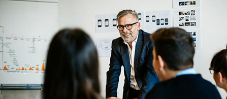A person with gray hair and glasses, wearing a dark blazer, sits smiling at the head of a table during a meeting. Two colleagues are seen from behind in the foreground, and a whiteboard with charts and sticky notes is on the wall behind.