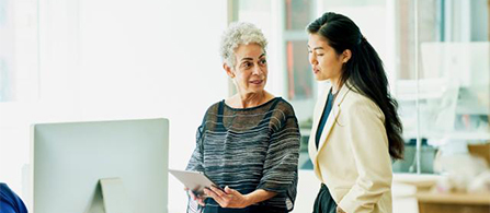 Two people stand together in a bright office looking at a tablet one of them holds. One has short gray hair and wears a textured gray top; the other wears a cream blazer over a dark top. Large windows brighten the background.