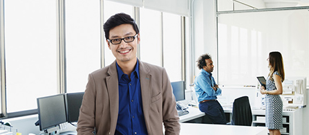 A person wearing glasses, a tan blazer, and a blue shirt stands smiling in a bright office. Two other people are blurred in the background near desks and large windows.