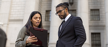 Two people stand outdoors in front of a large stone building, looking together at a tablet held by one of them. One wears a green jacket; the other wears a dark suit, white shirt, and tie, with a serious expression.