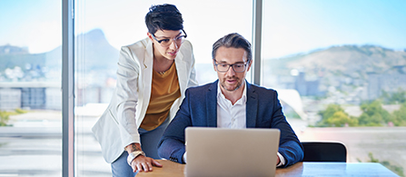 Two people look together at a laptop in a bright office with floor-to-ceiling windows overlooking a mountain landscape. One stands leaning in wearing a white blazer over an orange top; the other sits wearing a light shirt, both focused on the screen.