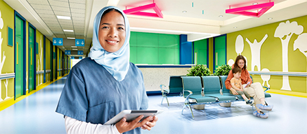 A person wearing a headscarf and scrubs stands in the foreground of a bright, colorfully decorated healthcare corridor, smiling and holding a tablet. A person sits in the waiting area behind them near cheerful wall murals and blue chairs.