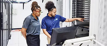 Two people in blue polo shirts stand together in a bright data center, looking at an open server rack. One points at the equipment while the other holds a clipboard and looks on. Rows of server cabinets fill the background.