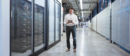 A person in a white button-down shirt and jeans walks down a wide corridor between server racks in a data center, looking down at a tablet with a relaxed expression.