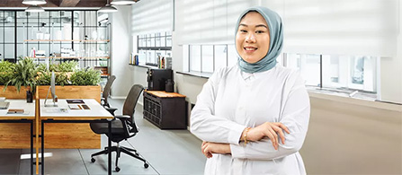 A person in a white blazer and light blue head covering stands with arms crossed and a smile in a modern open-plan office with desks, plants, and large windows behind them.