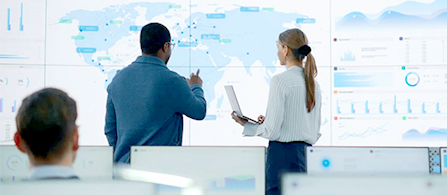 Two people stand facing large wall-mounted display screens showing a world map with data visualizations, charts, and graphs. One holds a laptop open. A third person is seated in the foreground with their back to the camera.