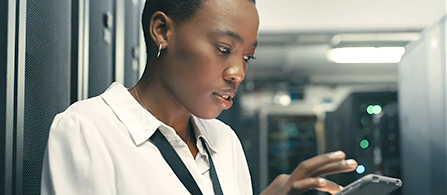 A person in a white button-down shirt stands in a server room, looking down at a smartphone with a focused expression. Server racks line the walls on both sides in the background.