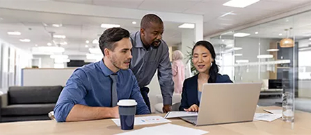 Three people are gathered around a laptop at a conference table in a bright, modern office. Two are seated and one stands leaning in, all focused on the laptop screen with engaged expressions.