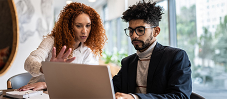 Two people work together at a laptop in a bright office. A person with curly red hair in a white top leans in and gestures while speaking, and a person in a dark blazer and glasses with a turtleneck looks at the laptop screen.