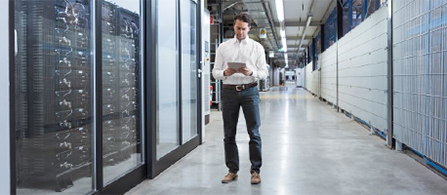 A person in a white shirt and dark trousers walks down a wide corridor between server racks in a data center, looking down at a tablet held in both hands.