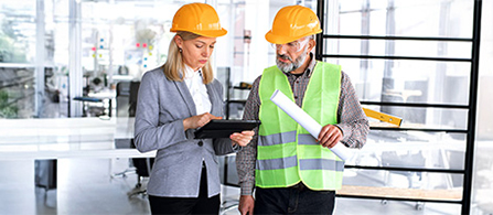 Two people wearing yellow hard hats stand in a bright industrial facility looking at a tablet together. One in a gray blazer holds the tablet, and the other, in a high-visibility safety vest, holds rolled documents and looks on.