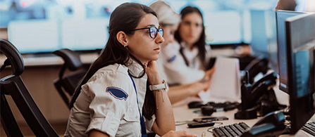 A person with dark hair and glasses, wearing a uniform shirt and lanyard, sits in profile at a workstation in a security operations center, resting a hand near their chin while focused on a monitor. Other people work at monitors in the background.