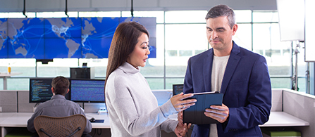 Two people stand together in a security operations center looking at a tablet one of them holds. One wears a gray sweater and the other a navy blazer. Large wall-mounted screens displaying a blue world map and a person working at monitors are visible in the background.