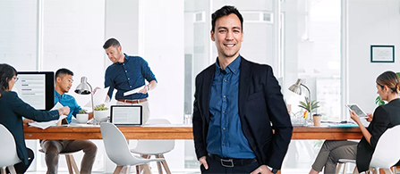 A person in a dark blazer and blue shirt stands with hands in pockets and a slight smile in a bright, modern office. Several other people are working at desks with laptops and monitors in the background.