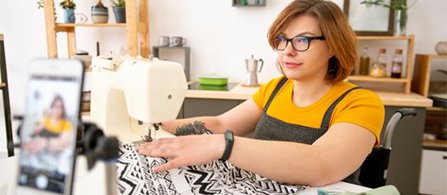 A person wearing glasses, a yellow top, and a dark apron sits at a sewing machine working with patterned fabric, with a smartphone propped on a stand in the foreground as if recording or streaming in a home studio setting.
