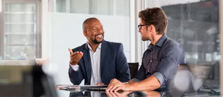 Two people seated across from each other at a small round table near large windows in a bright office, smiling and engaged in a friendly conversation.