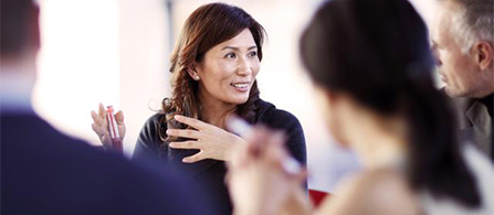 A person with long dark hair speaks animatedly with both hands raised in a small group discussion, smiling, with other participants visible in the foreground and background in soft focus.