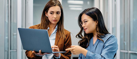 Two people stand close together in an office corridor looking at a laptop held by one of them. One wears a brown blazer over a light top; the other wears a light blue blouse and glasses and points at the screen. Both have focused expressions, with glass-walled offices blurred behind them.