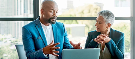 Two people sit at a table in conversation near large windows. One wears a blue blazer and gestures with both hands while speaking; the other wears a dark blazer with hands clasped on the table, listening and smiling.