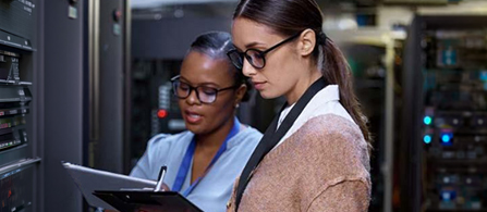 Two people work side by side in a data center aisle lined with server racks. One holds a clipboard and writes; both wear glasses and look focused on the equipment.