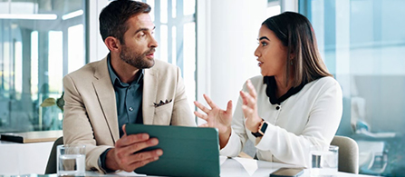 Two people sit at a table in a bright office having a discussion. One wears a tan blazer and holds a tablet; the other wears a white blazer and gestures while speaking.