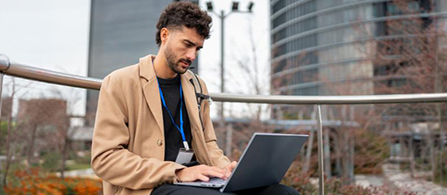 A person in a tan jacket wearing a lanyard sits outdoors on a railing-lined terrace using a laptop, with an office building and greenery blurred behind them.