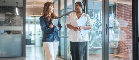 Two people walk and talk together down an office hallway, each carrying documents, with glass partitions along the corridor.